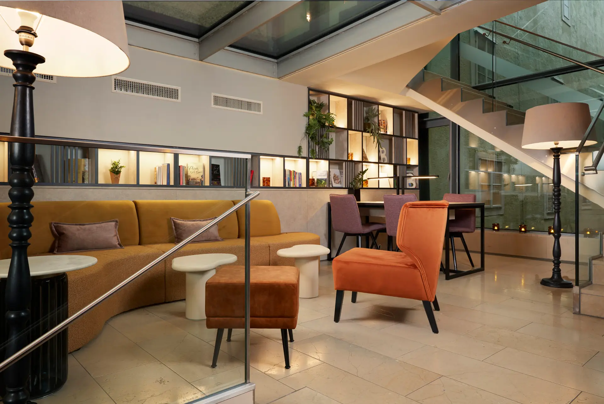 Hotellobby mit geschwungenem ockerfarbenem Sofa, Samtsesseln, Bücherregalen und Glastreppe
