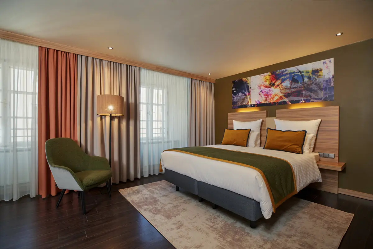 Hotelzimmer mit Doppelbett, grünem Polstersessel, Stehlampe und großformatigem Kunstwerk über dem Kopfteil, abendlich warm beleuchtet