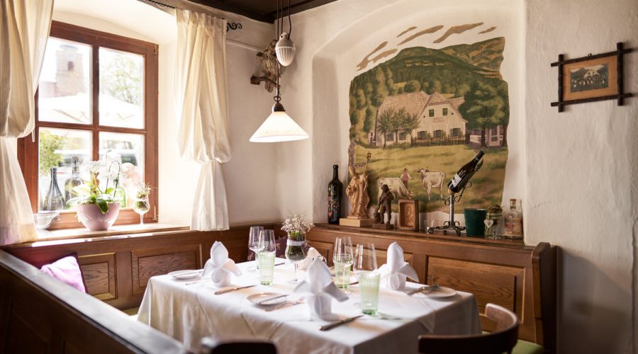 Eingedeckter Ecktisch in einer Stube am Fenster, daneben eine Wandmalerei mit Bauernhof und Kühen